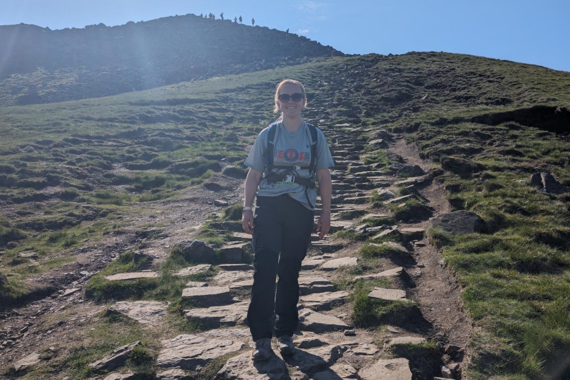 A woman stands confidently on a rocky path, embodying the spirit of a Games Workshop volunteer in nature.